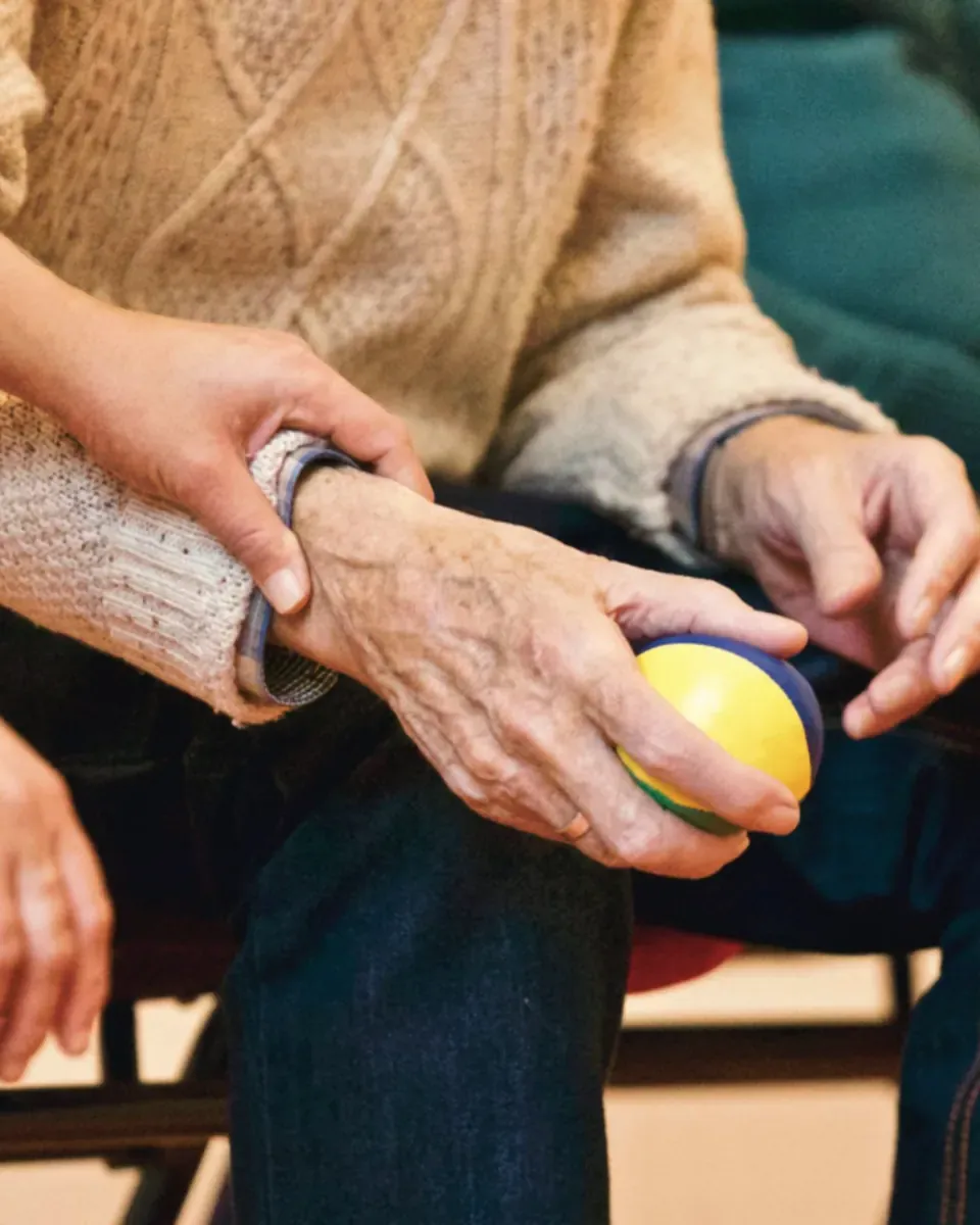 Nahaufnahme: Eine ältere Person hält einen bunten Therapieball in der Hand, eine zweite Hand stützt behutsam ihr Handgelenk.
