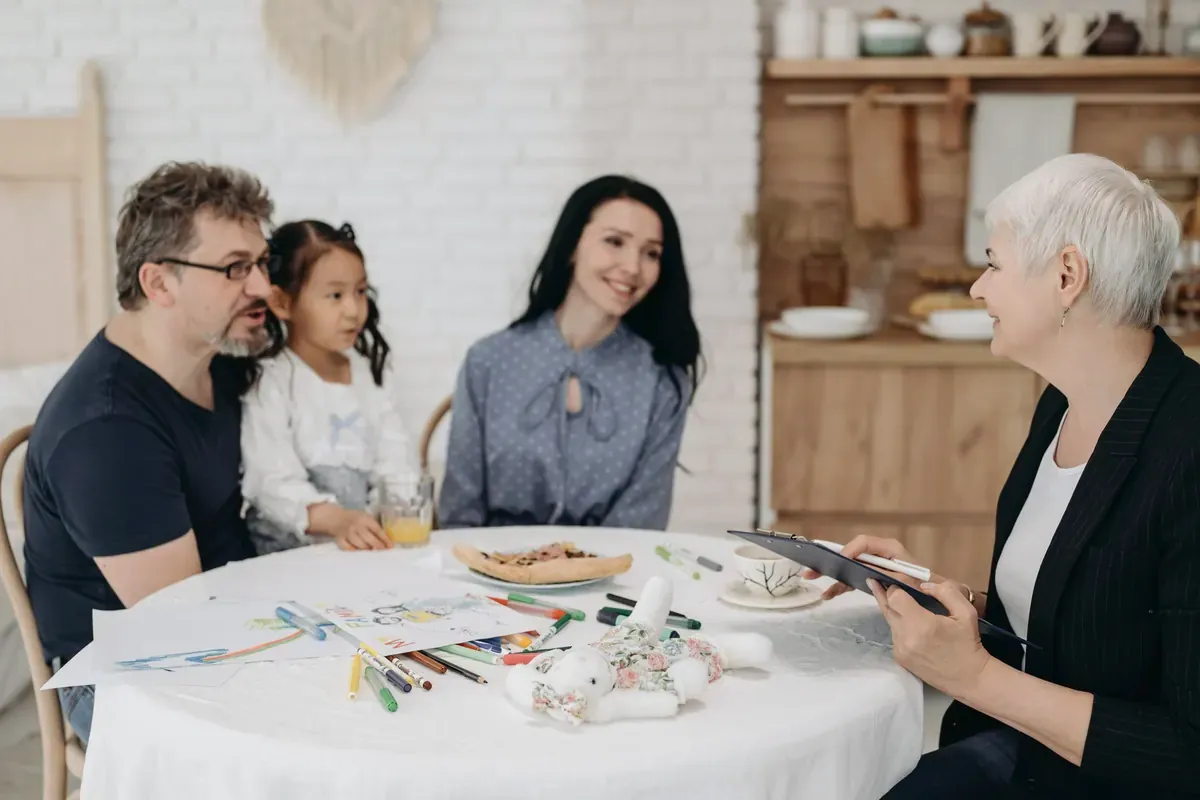 Eine Familie mit Vater, Mutter und kleiner Tochter sitzt an einem runden Tisch mit Kinderzeichnungen und Stiften und spricht mit einer Fachfrau, die ein Klemmbrett hält.