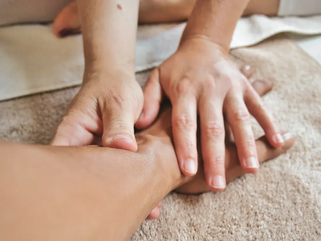 Nahaufnahme: Zwei Hände massieren behutsam die Hand und das Handgelenk einer anderen Person, die auf einem hellbraunen Frotteetuch liegt.