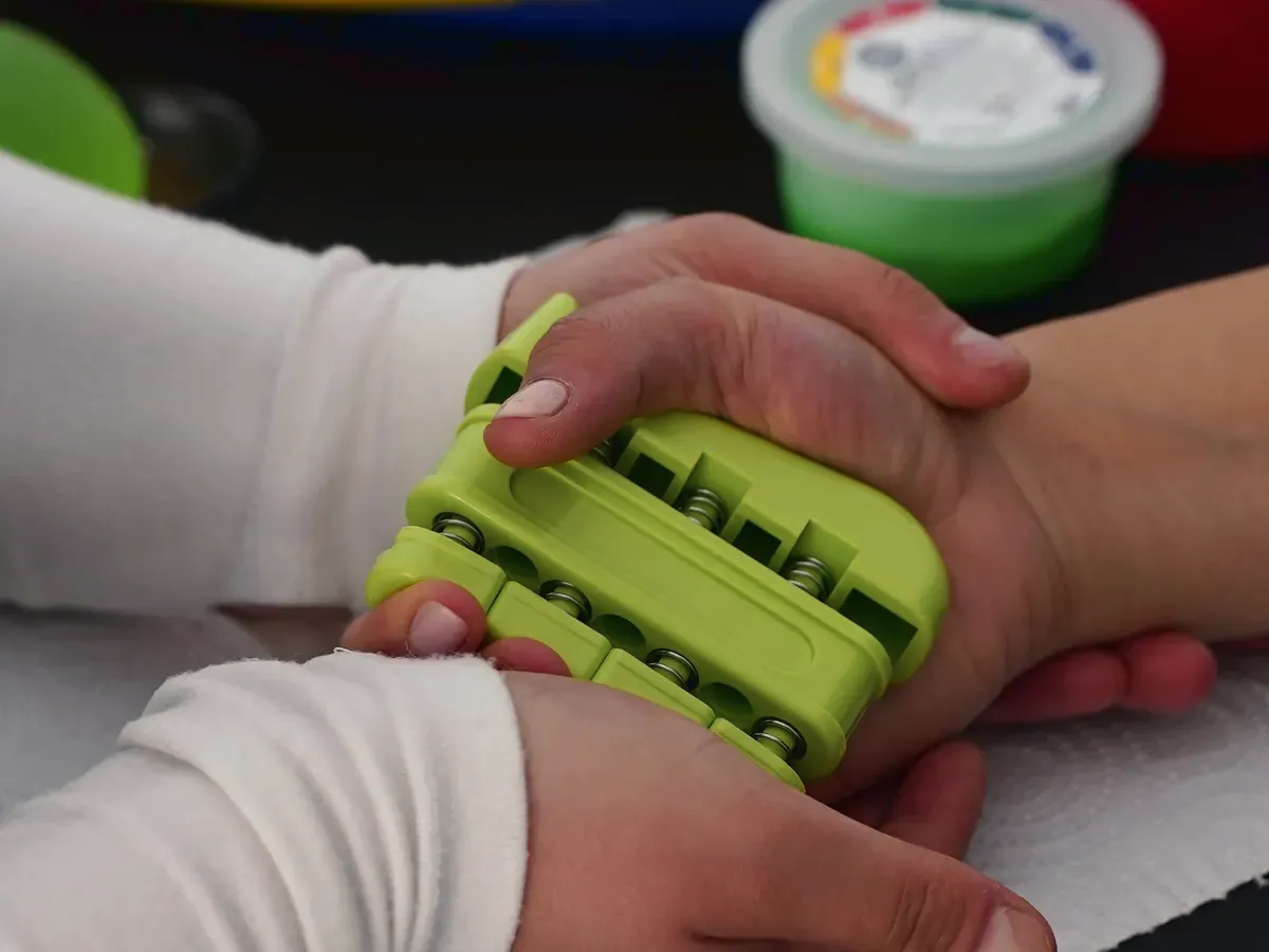 Nahaufnahme am Therapietisch: Zwei Hände in weißen Ärmeln führen eine Hand an einem grünen Fingertrainer, im Hintergrund steht eine Dose Therapieknete.