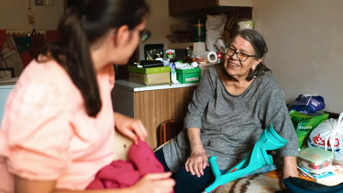 Eine ältere Frau mit Unterarmgehstützen sitzt lachend in ihrer Wohnung. Ihr gegenüber sitzt eine junge Therapeutin und hält ein pinkfarbenes Kleidungsstück, das die beiden gemeinsam sortieren.