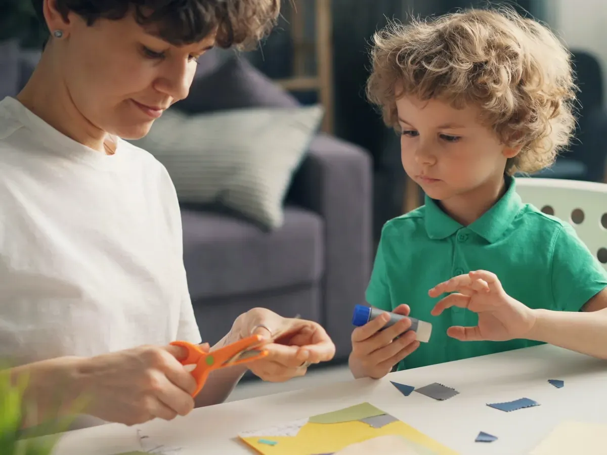 Eine Frau schneidet mit einer orangefarbenen Kinderschere Papier zu, neben ihr sitzt ein Junge im grünen Poloshirt mit einem Klebestift am Basteltisch.