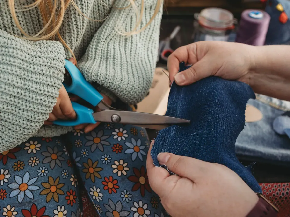 Ein Kind schneidet mit einer blauen Schere Jeansstoff zu, eine erwachsene Person hält den Stoff straff.