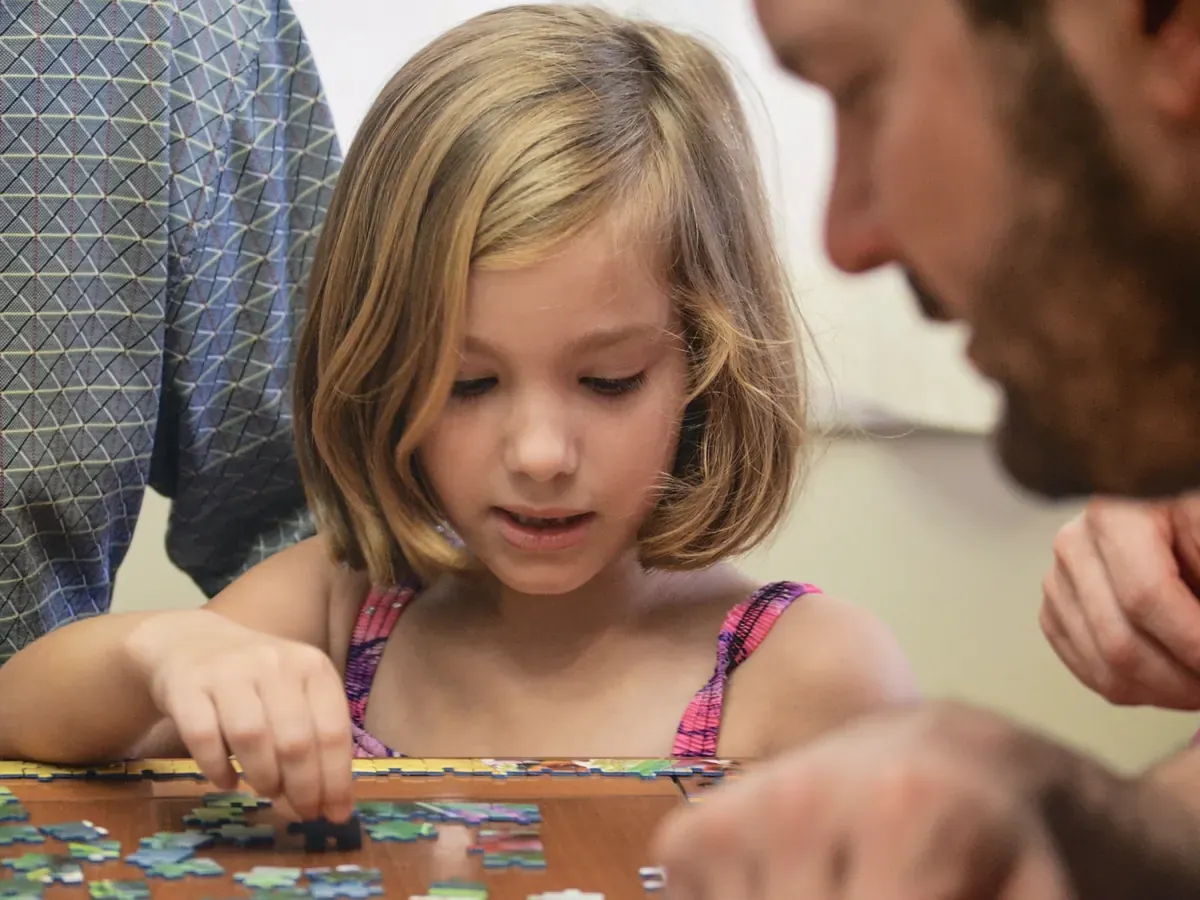 Ein Mädchen legt am Tisch ein Puzzle, ein Mann beugt sich zu ihr und puzzelt mit.